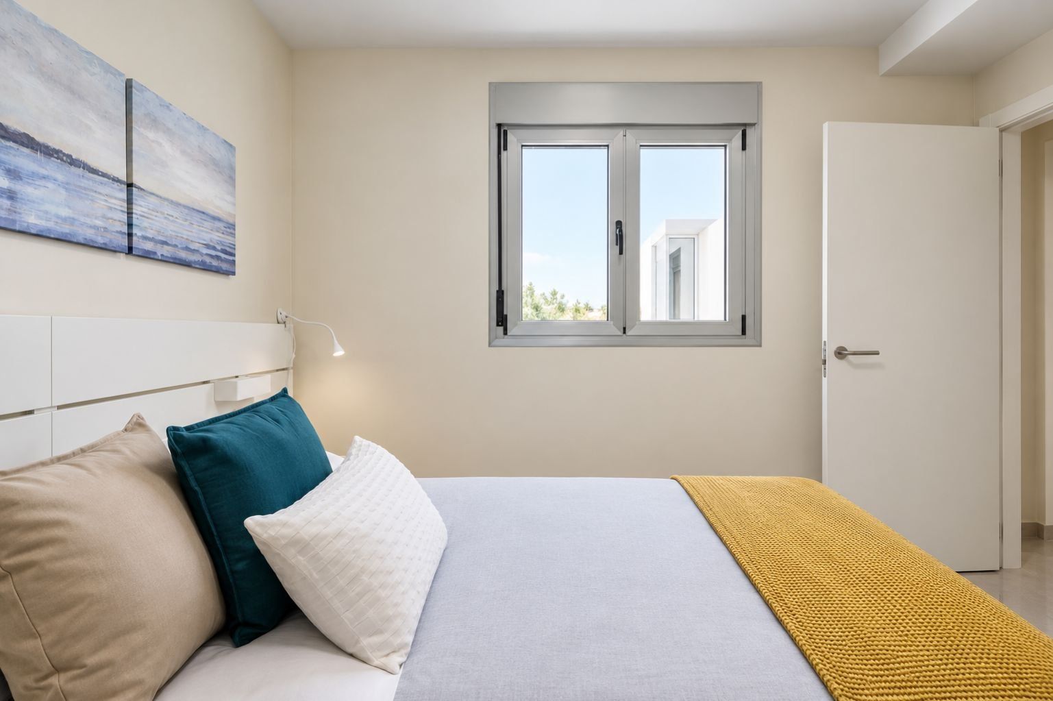 A bedroom with a bed, pillows, a bedside lamp, a window, and a door.