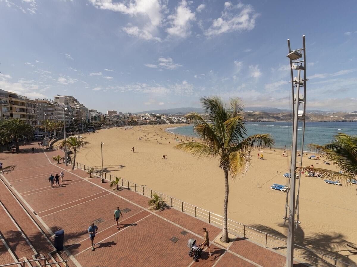 A beach with a paved walkway, palm trees, and a view of the sea and distant buildings.