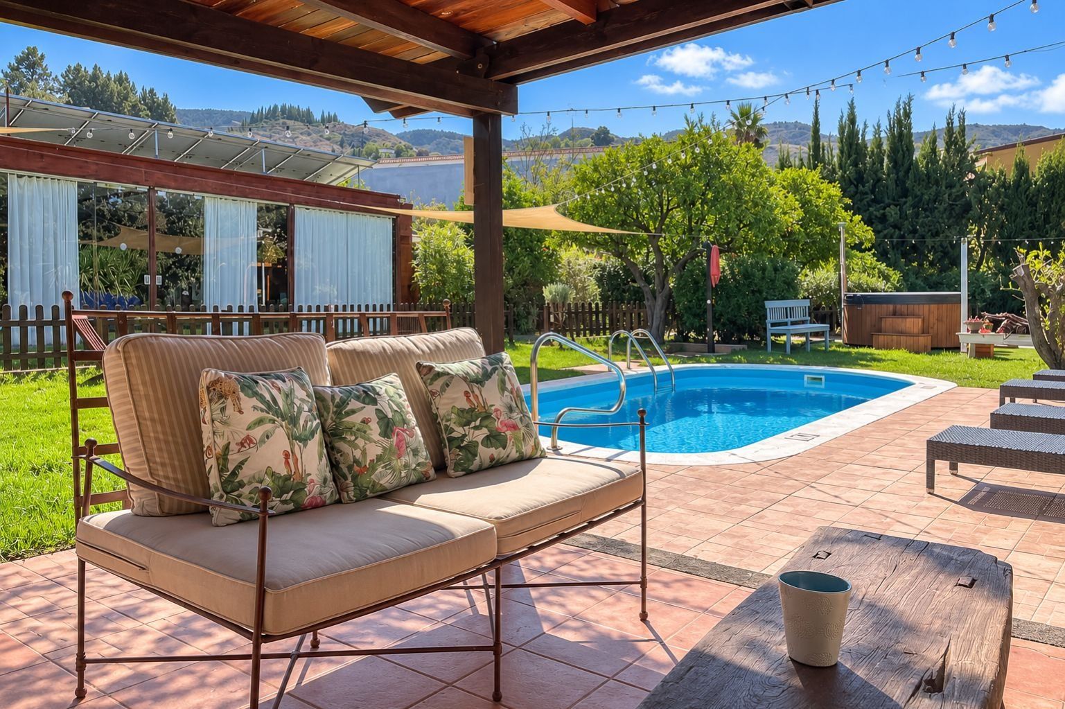 Un espace extérieur abrité comprenant une piscine, un banc en bois et un coin salon avec des coussins à motifs floraux.