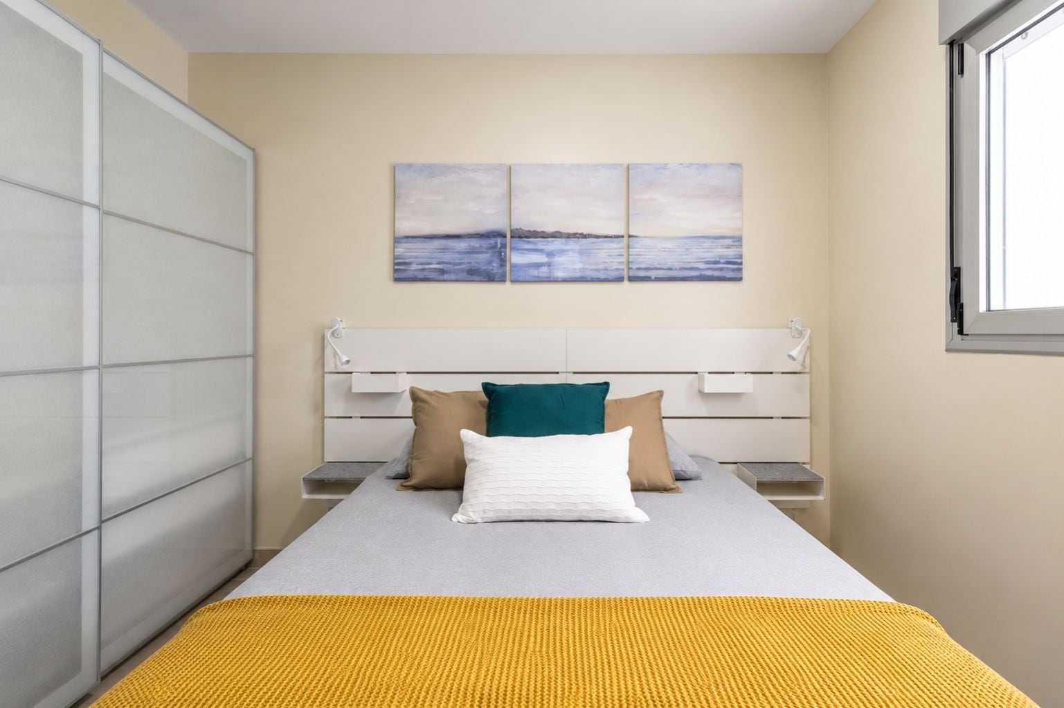 Ein Schlafzimmer mit Bett, Kissen, Nachttisch, Fenster und einem Wandbild.