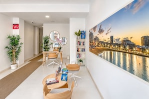 A modern hotel room with a cityscape view, a dining area with a table and chairs, and a bookshelf with books. - GuestReady - 03B Livian Suites Int 4 pax (Las Palmas de Gran Canaria)