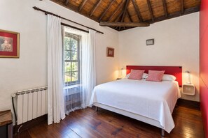 Une chambre avec un plancher en bois, un lit avec une literie blanche, une tête de lit rouge et une fenêtre avec des rideaux blancs.