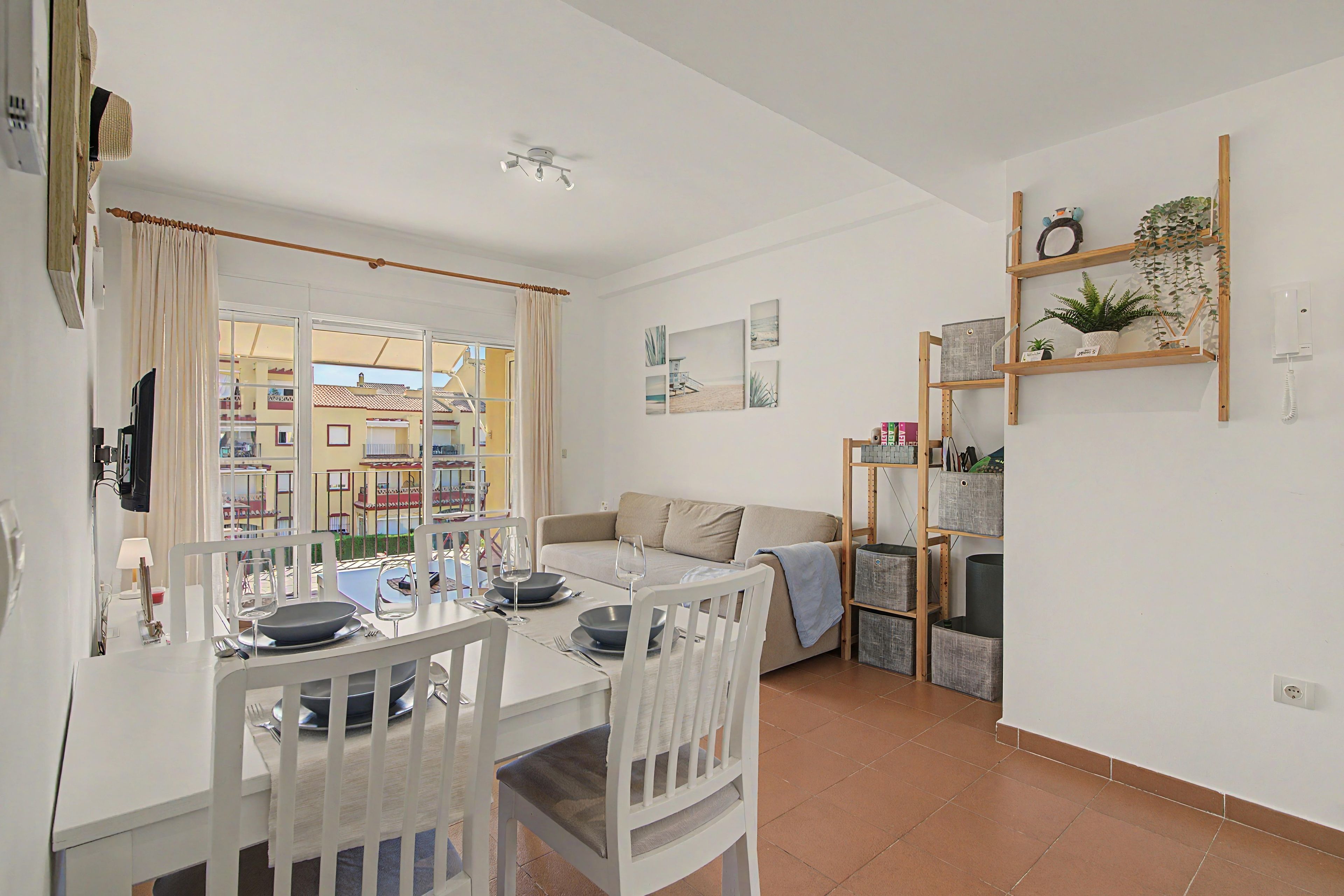 A dining area with a table set for two, a sofa, and a bookshelf with plants.