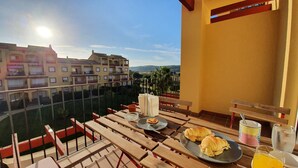 Un balcon avec une table dressée pour un repas, comprenant une assiette avec de la nourriture et des boissons, et une vue sur un ensemble d’immeubles.