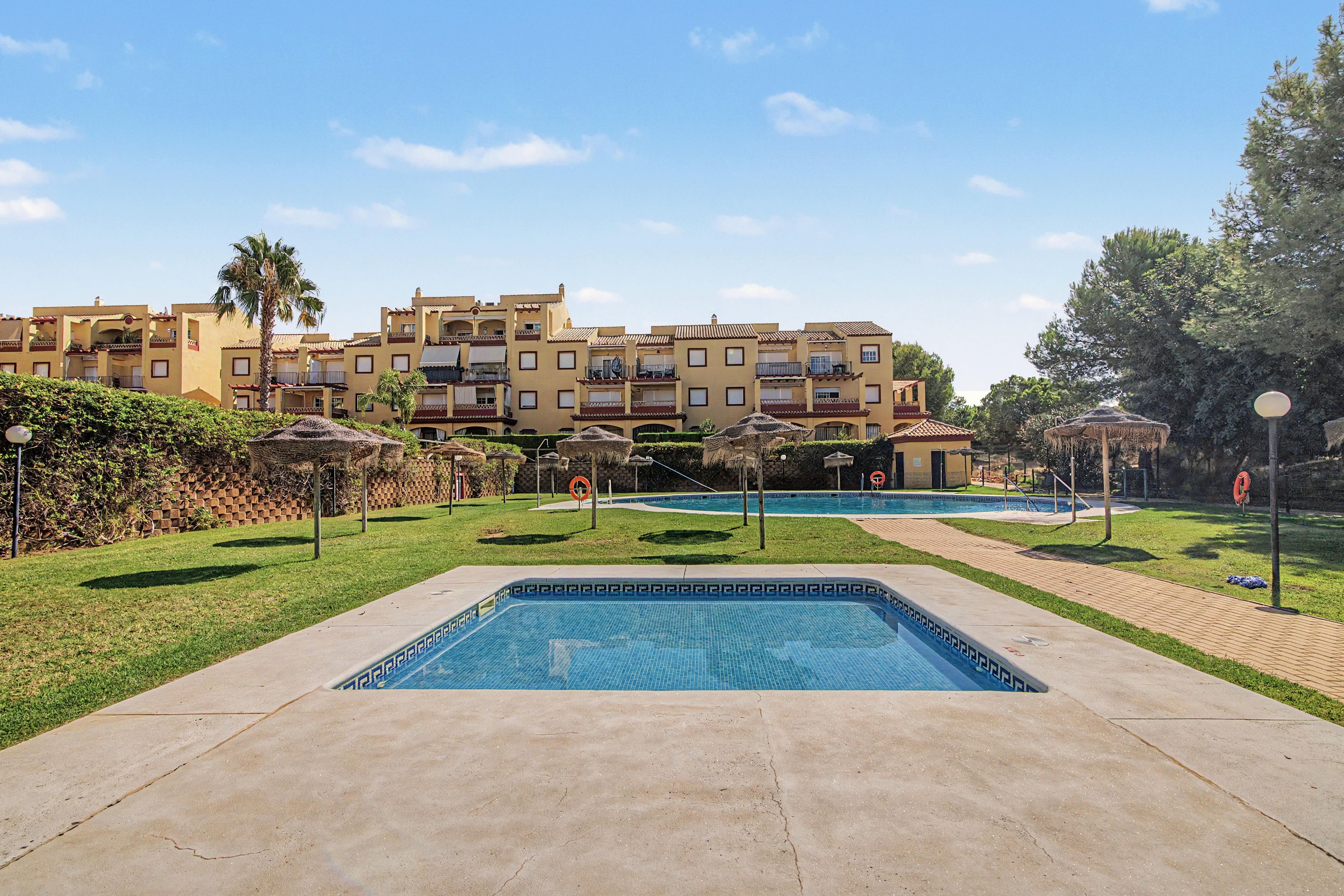 A swimming pool area with a clear sky, a building with balconies, and a grassy lawn.