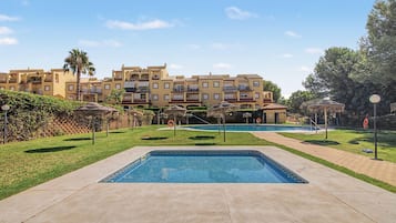 A swimming pool area with a clear sky, a building with balconies, and a grassy lawn.