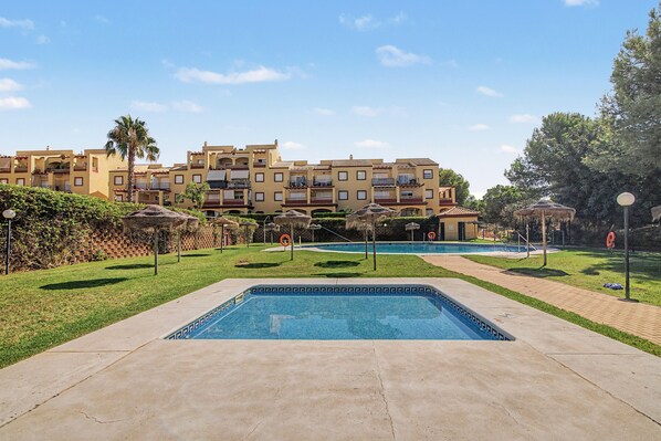 A swimming pool area with a clear sky, a building with balconies, and a grassy lawn.