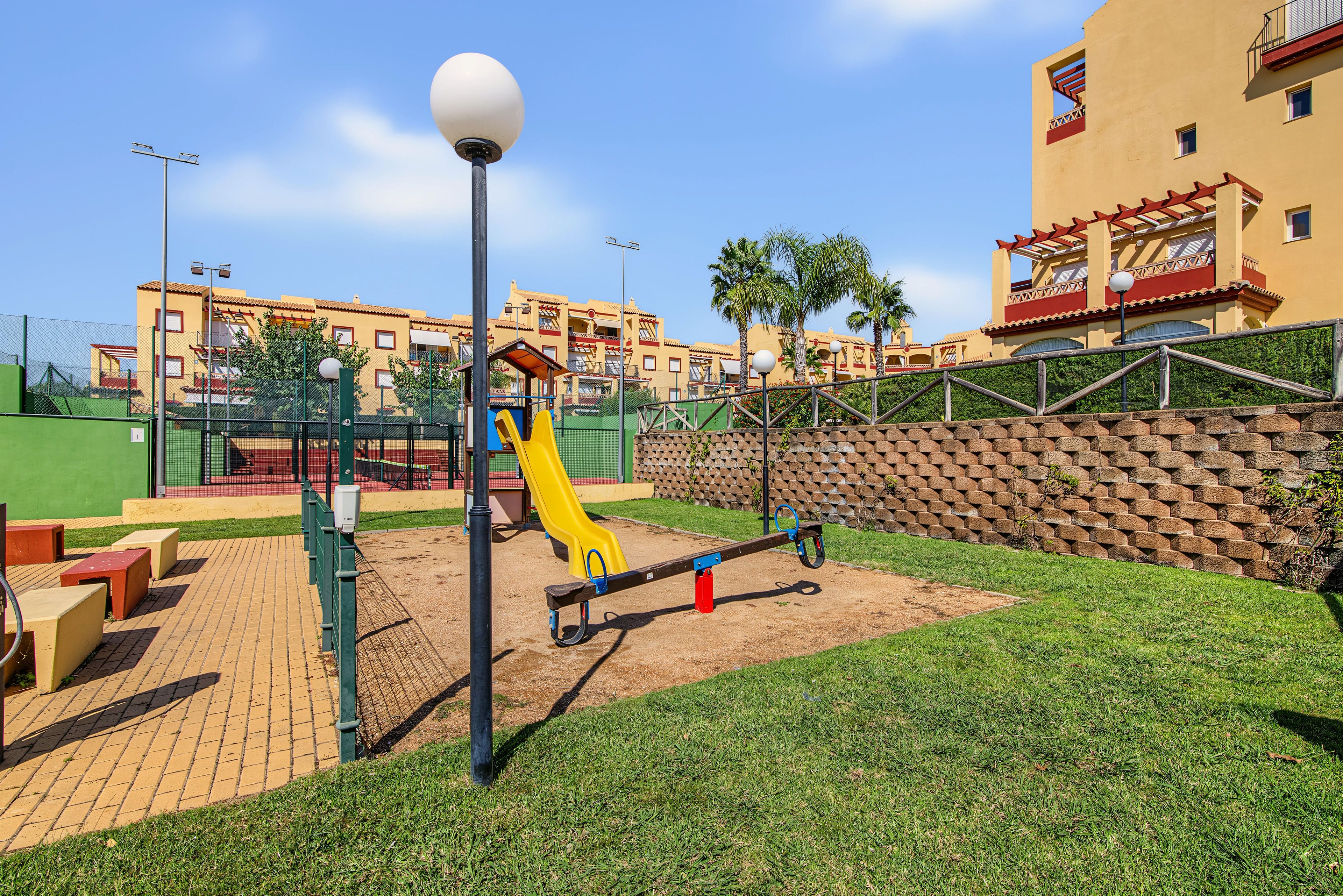 Un parque infantil con un sube y baja, un tobogán y una estructura para escalar, rodeado por un muro de ladrillo y un edificio con balcones.