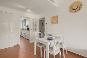 Uma área de refeições com uma mesa branca posta para quatro pessoas, uma cozinha ao fundo e um chapéu de palha com a inscrição 'hello sunshine' na parede.