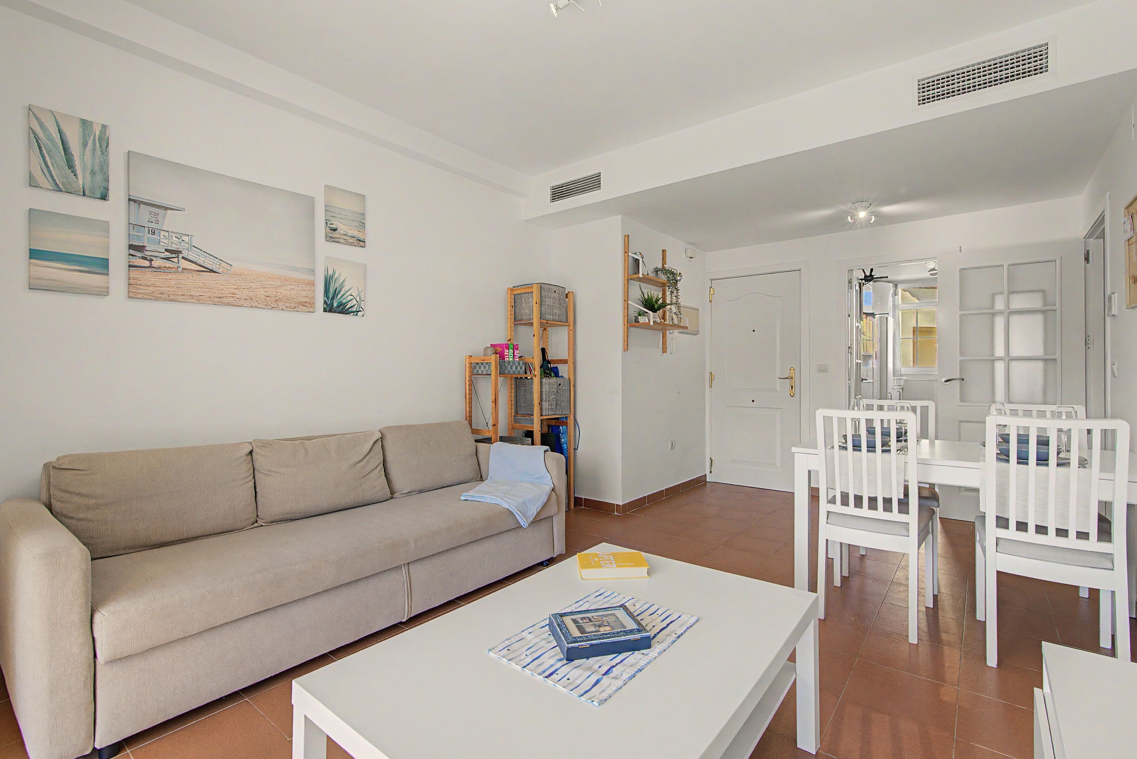 A living room with a beige sofa, a white coffee table with a book, a dining area with a white table and chairs, and a kitchen visible through an open door.