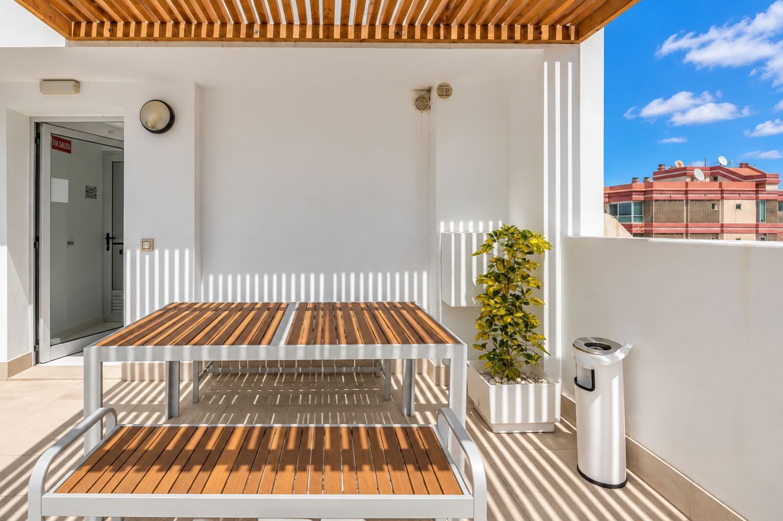 Un balcón con mesa y sillas de madera, una planta en maceta y un cubo de basura.