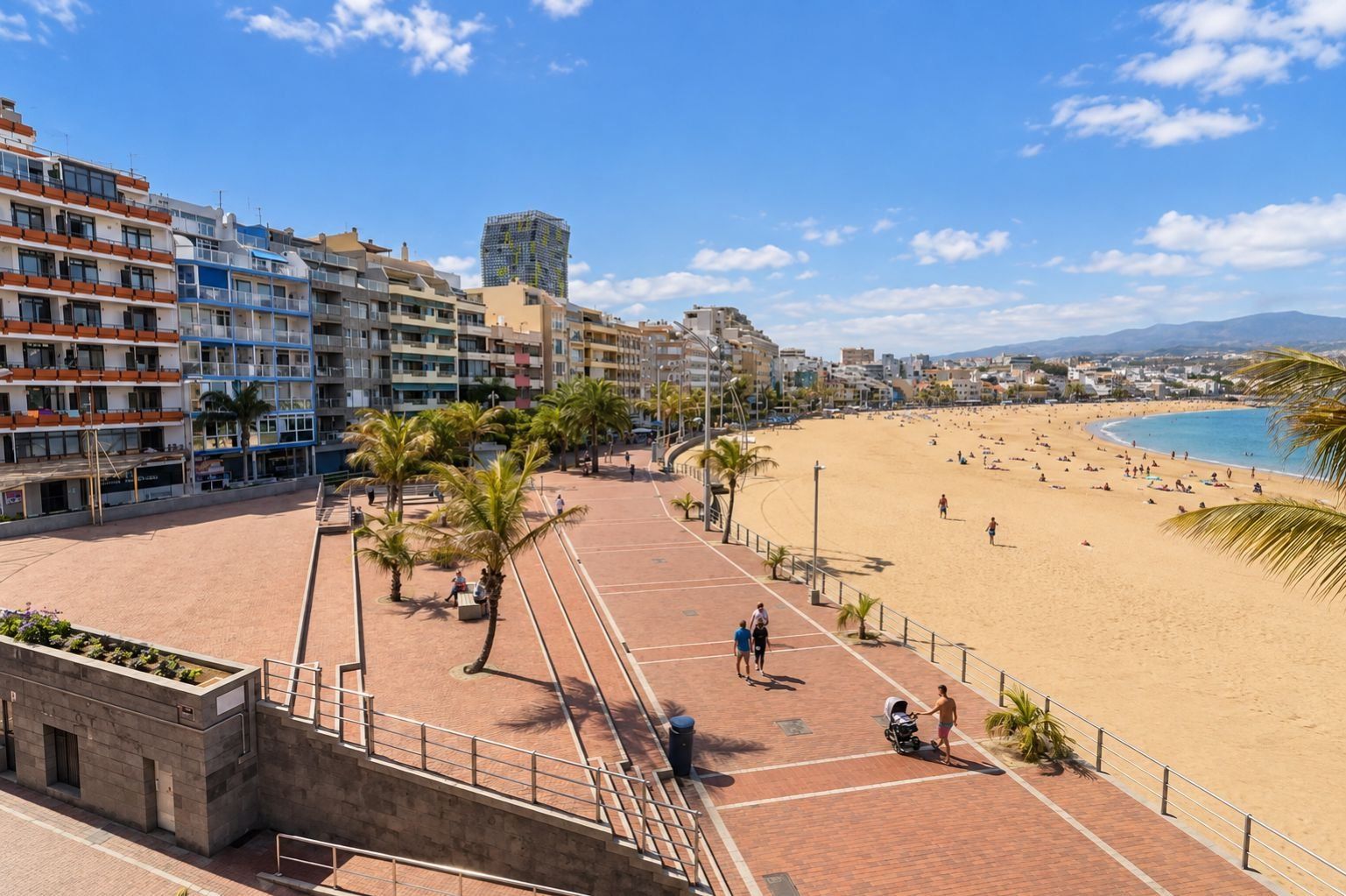 Une promenade en bord de mer, avec une plage de sable, des palmiers et une rangée d’immeubles à plusieurs étages.