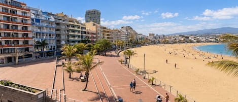 A beachside promenade with a sandy beach, palm trees, and a row of multi-story buildings.