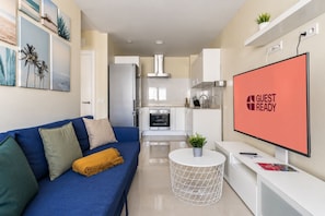 A modern living room with a blue sofa, a flat-screen TV displaying 'GUEST READY,' and a kitchen in the background. - GuestReady - 04A Livian Suites Int 3 Pax/Terraza (Las Palmas de Gran Canaria)