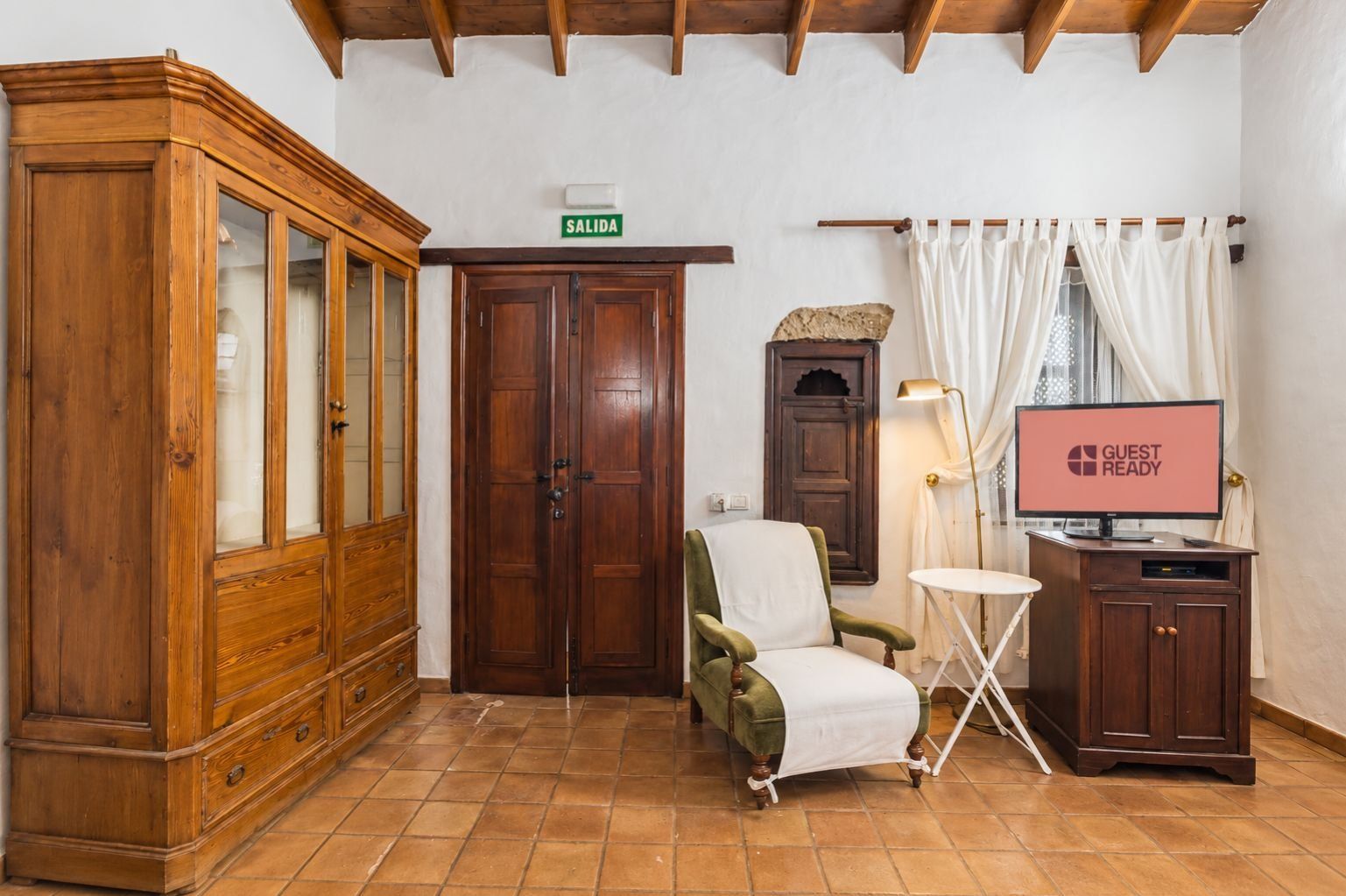 A room with a wooden cabinet, a green armchair, a television displaying 'GUEST READY', and a sign indicating 'SALIDA'.