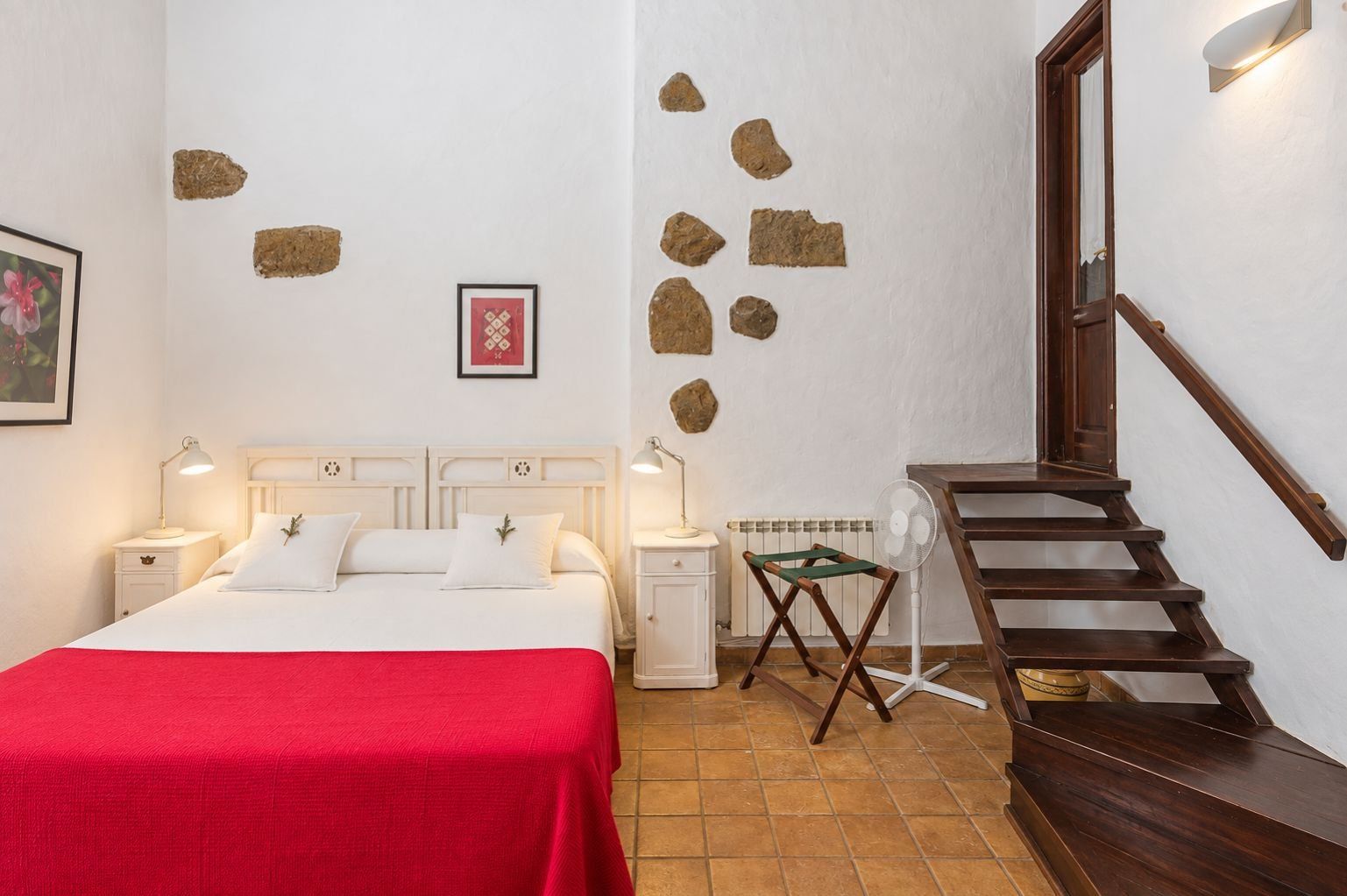 Habitación de hotel con una cama, mesitas de noche, una colcha roja, una escalera de madera y una pared con piedras decorativas.
