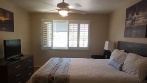 A bedroom with a bed, a ceiling fan, a window with blinds, a TV on a dresser, and artwork on the walls. - Machronicles Properties at Sand Pebbles Resort in beautiful Solana Beach (Solana Beach)
