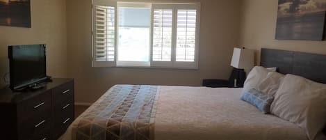 A bedroom with a bed, a ceiling fan, a window with blinds, a TV on a dresser, and artwork on the walls.