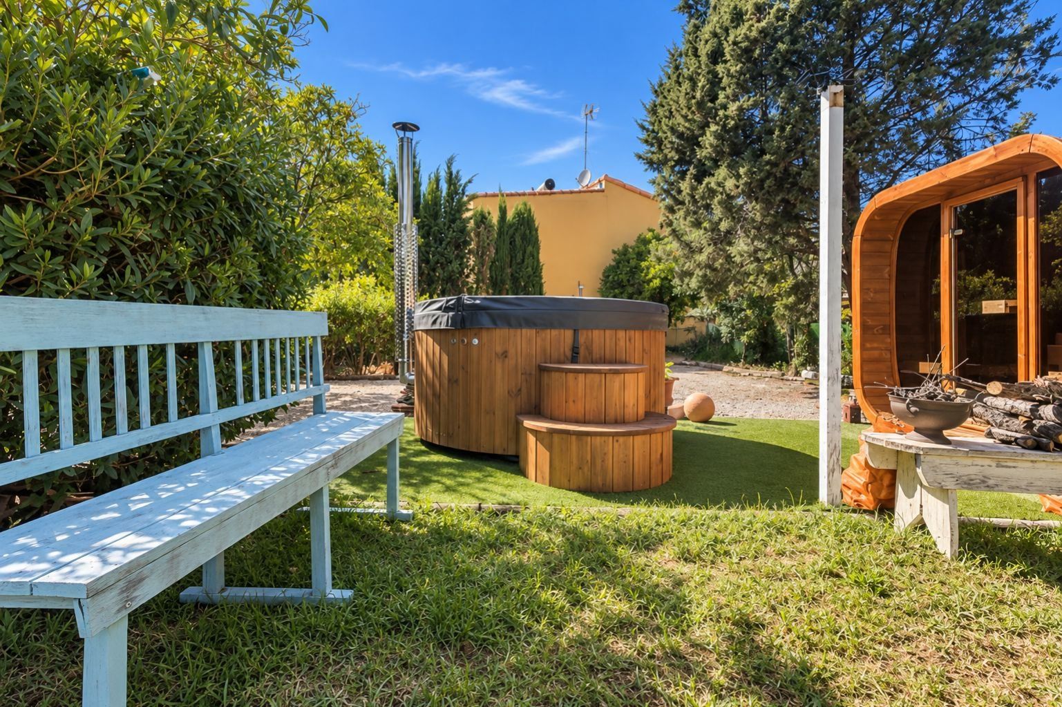 A wooden bench, a hot tub, and a wooden structure with a fire pit in a garden.