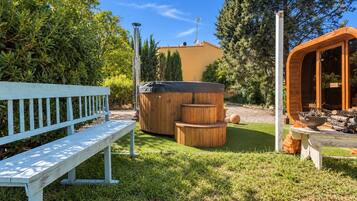 A wooden bench, a hot tub, and a wooden structure with a fire pit in a garden.