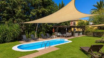 Una piscina con zona sombreada, sillas de descanso y un porche cubierto.