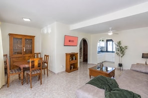 A spacious living room with a dining area, a kitchen visible through an archway, and a flat-screen TV mounted on the wall.