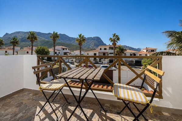 Uma varanda com mesa de madeira e duas cadeiras, com vista para uma área residencial com palmeiras e casas.