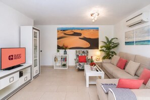 A modern living room with a flat-screen TV, a bookshelf, a sofa, a coffee table, and a painting of a desert landscape. - GuestReady - Sun Club Terrace and Pool (San Bartolomé de Tirajana)