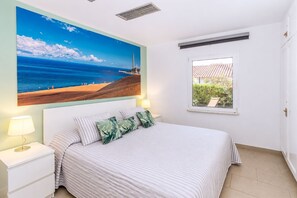 Ein Schlafzimmer mit einem Bett, einem Nachttisch, einer Lampe, einem Fenster mit Blick auf ein Gebäude und einem Gemälde mit Strandmotiv.