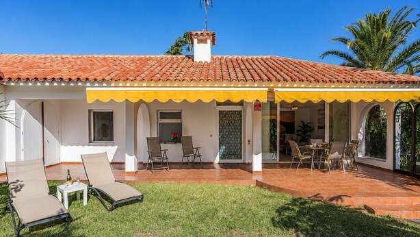 Uma casa com telhado de cerâmica, um alpendre coberto com cadeiras e um gramado.