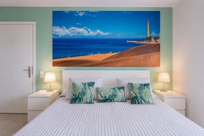 A bedroom with a large bed, two bedside lamps, a nightstand, and a painting of a beach scene. - GuestReady - Sun Club Terrace and Pool (San Bartolomé de Tirajana)