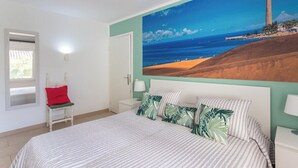 Um quarto com uma cama, uma mesinha de cabeceira, uma cadeira, um quadro com uma paisagem costeira e uma janela.