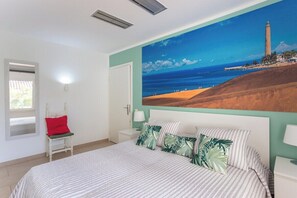 A bedroom with a bed, a nightstand, a chair, a painting of a coastal scene, and a window. - GuestReady - Sun Club Terrace and Pool (San Bartolomé de Tirajana)