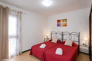 A double bed with two pillows and two folded towels, a nightstand with a lamp, a painting on the wall, and a window with curtains. - GuestReady - Bello Horizonte Sea View and Pool (San Bartolomé de Tirajana)