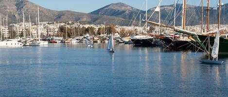 Un puerto deportivo con veleros, un velero con una persona a bordo, y un paisaje urbano con montañas al fondo.
