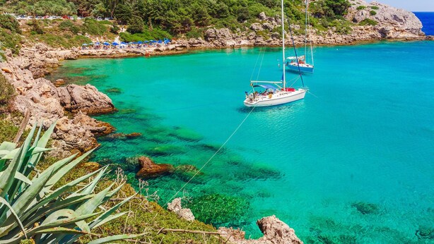 A coastal area with clear turquoise waters, rocky terrain, and a sailboat anchored near the shore. - Sun-Kissed Sailing Adventure in the Dodecanese Islands (Rodos)