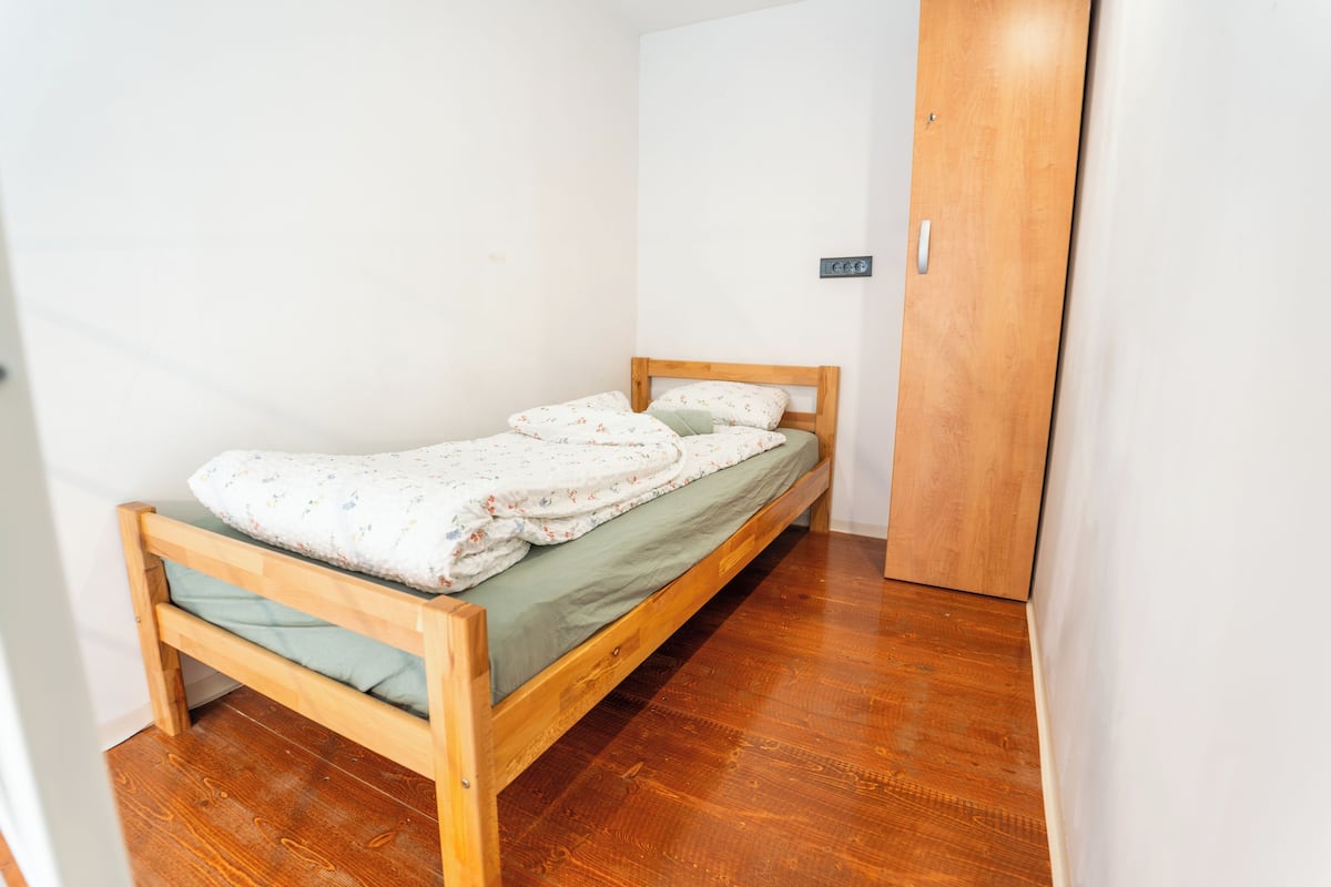 Una cama de madera individual con un colchón verde, una manta blanca y una almohada estampada.