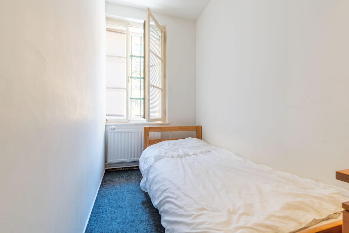Una habitación pequeña y estrecha con una cama individual, una ventana con contraventanas y un radiador.