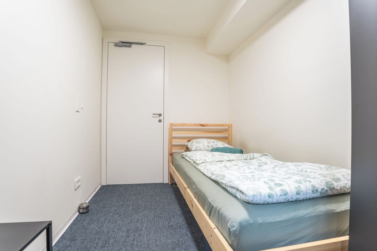 Una habitación pequeña con una cama individual, cabecera de madera, alfombra gris y una puerta blanca.