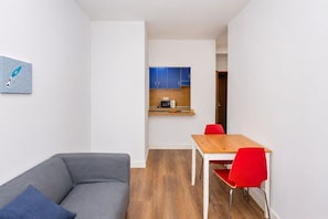 Un espacio habitable compacto con un sofá gris, una mesa de comedor de madera con sillas rojas, una kitchenette con armarios azules y una obra de arte abstracta colgada en la pared.
