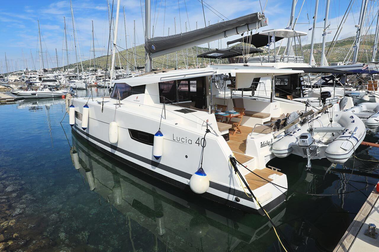 Un puerto deportivo con varios veleros amarrados, un yate blanco llamado 'Lucia 40' con cubierta y zona de estar techada, y una lancha inflable más pequeña.