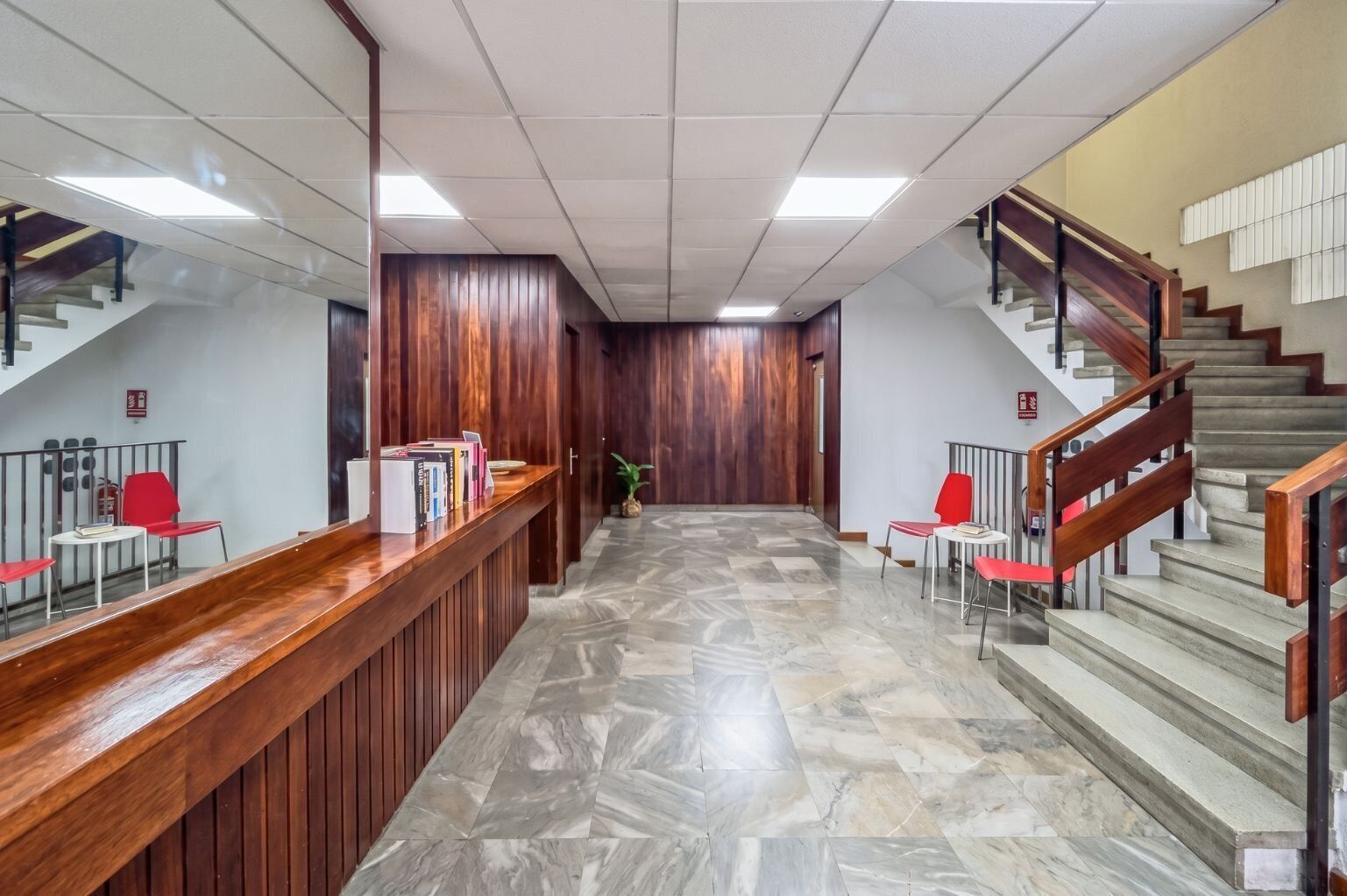 Zona de recepción con mostrador de madera, sillas rojas, suelo de mármol y una escalera con barandillas de madera.
