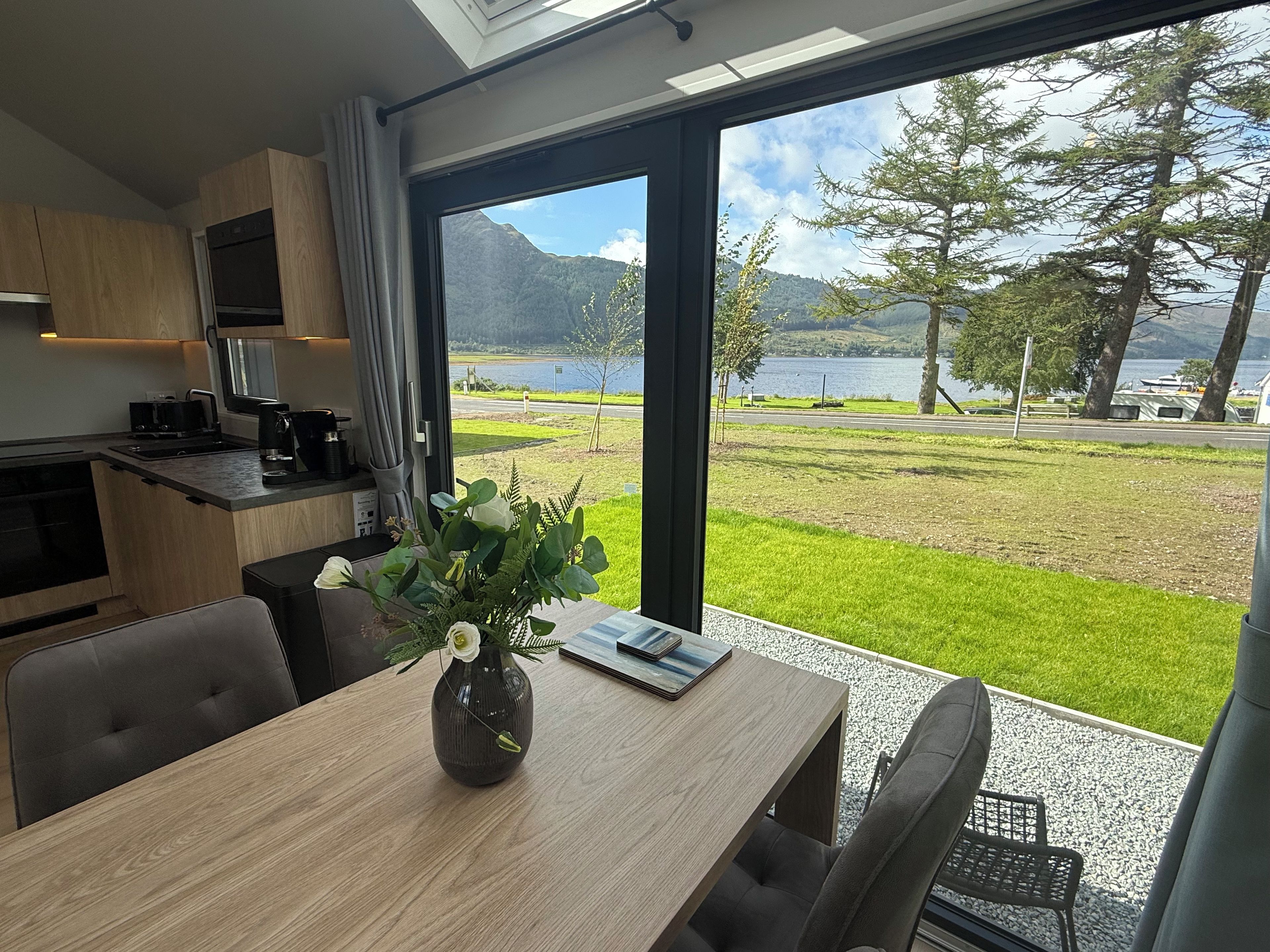 A modern kitchen with a large window, a dining table with a vase, and a view of a lake and mountains.