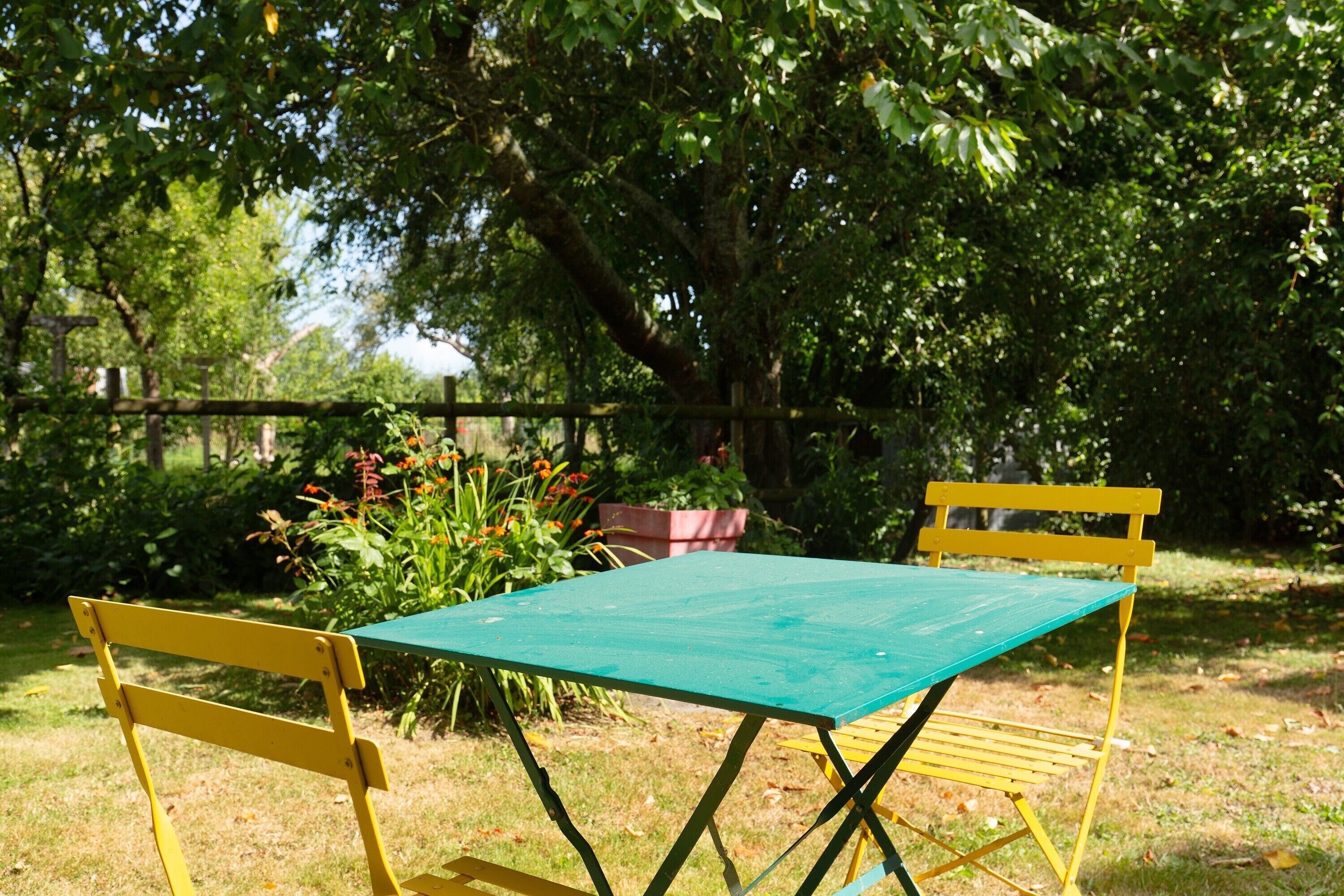 A green picnic table with yellow chairs in a grassy outdoor area with trees and flowers.