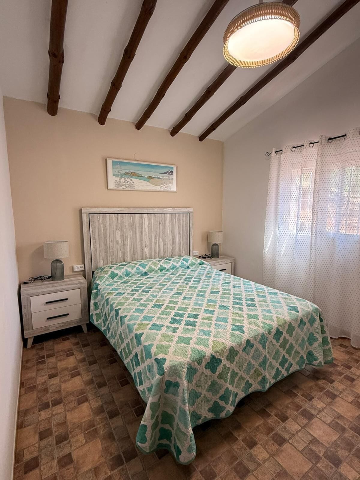 A bedroom with a bed, a nightstand, a lamp, a painting on the wall, and a window with curtains.