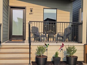 A residential building with a number '49F' on the wall, a front door, a small porch with a railing, two chairs, and three potted plants.