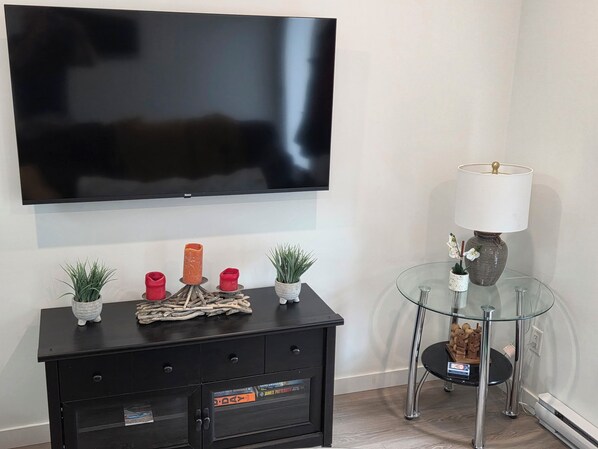Uma sala de estar com televisão de tela plana na parede, um móvel preto para entretenimento com objetos decorativos, uma mesa de centro de vidro com abajur e uma pequena planta em vaso.