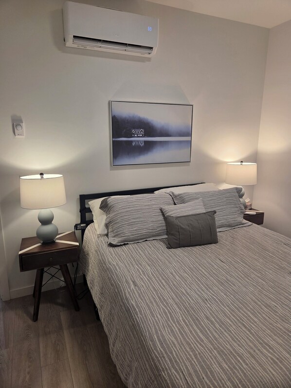 A bedroom with a bed, two bedside lamps, a wall-mounted air conditioner, and a framed picture. - Stylish pet-friendly 1-bedroom home on the border of Kelowna/Lake Country (Kelowna)