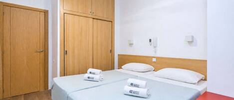 A hotel room with two beds, wooden wardrobe, a red side table, and a ceiling fan.