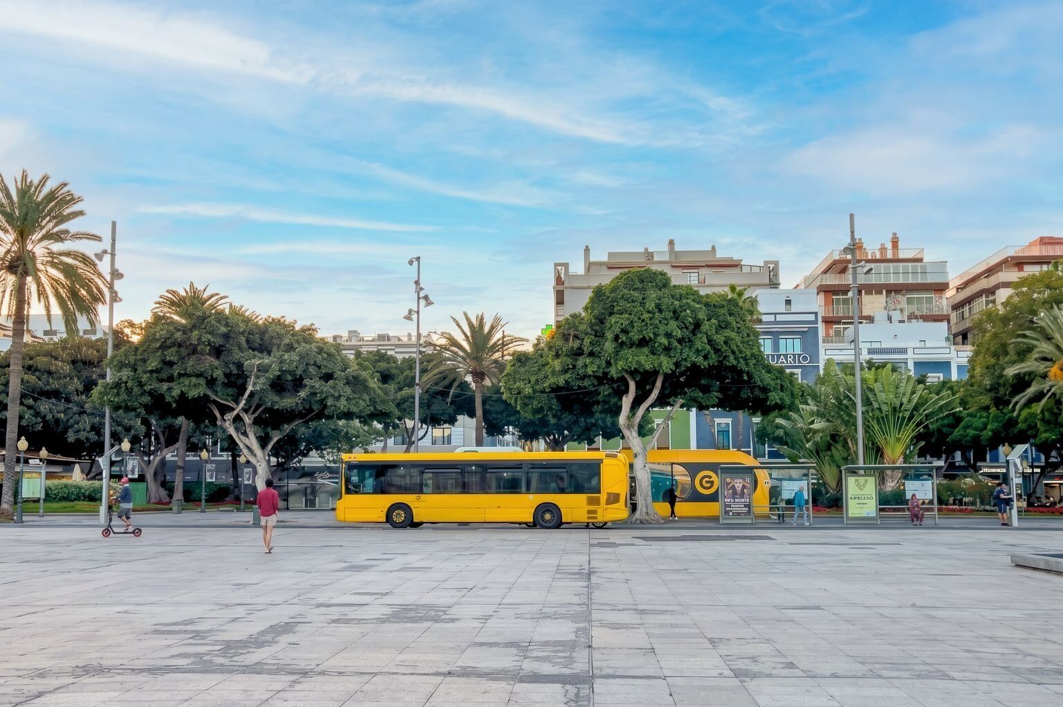A spacious public square with a yellow bus, palm trees, and modern buildings in the background.